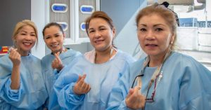 Four Dental Assistants in disposable isolation gowns smiling and making Korean finger hearts.
